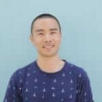 A man in a blue shirt posing happily against a blue wall.