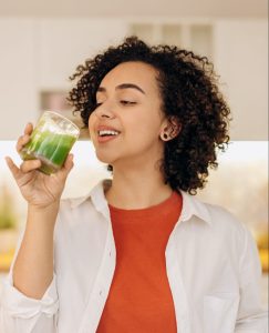 A woman sipping on a glass of green juice with matcha syrup in the kitchen.