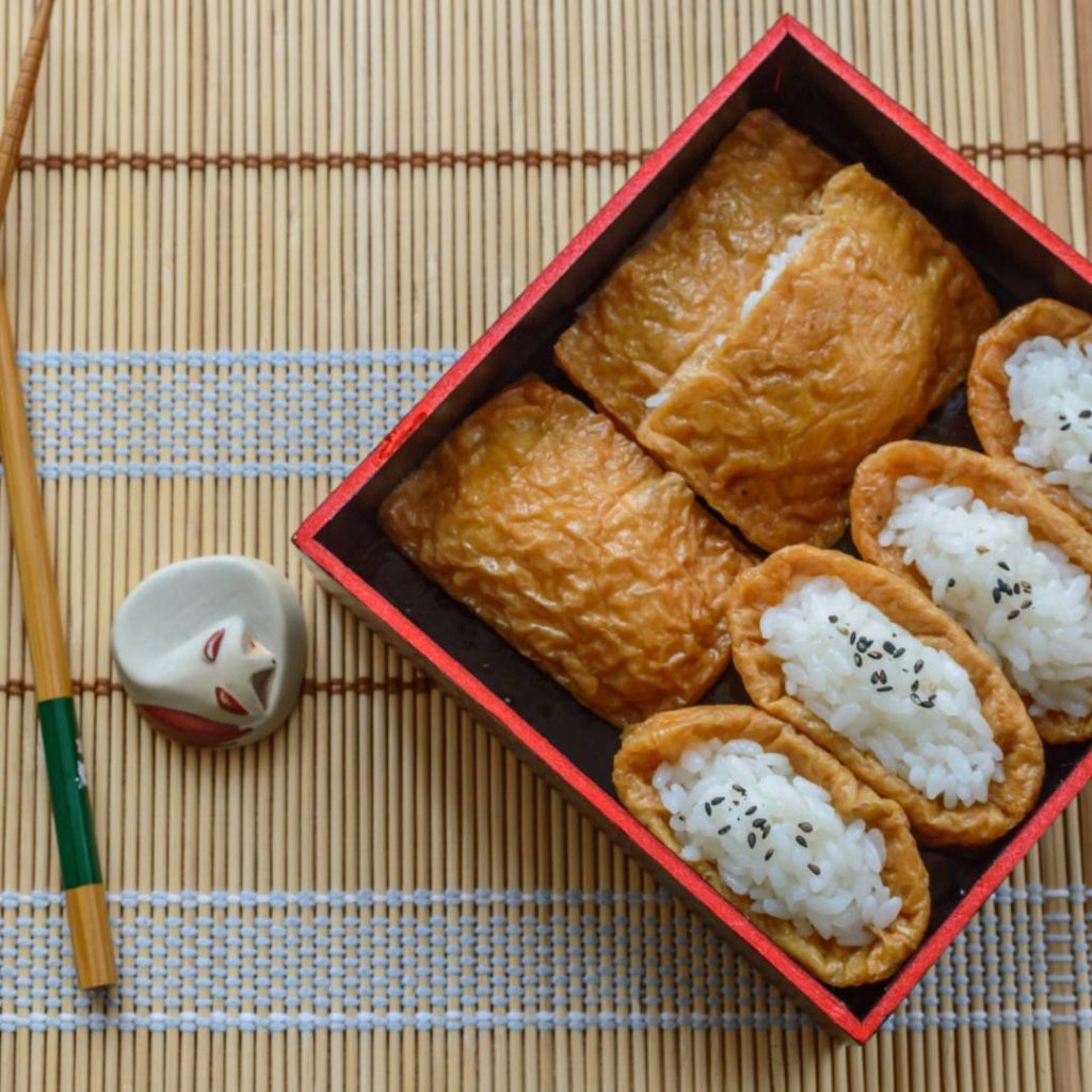 Japanese food in a box with chopsticks, featuring inari sushi.