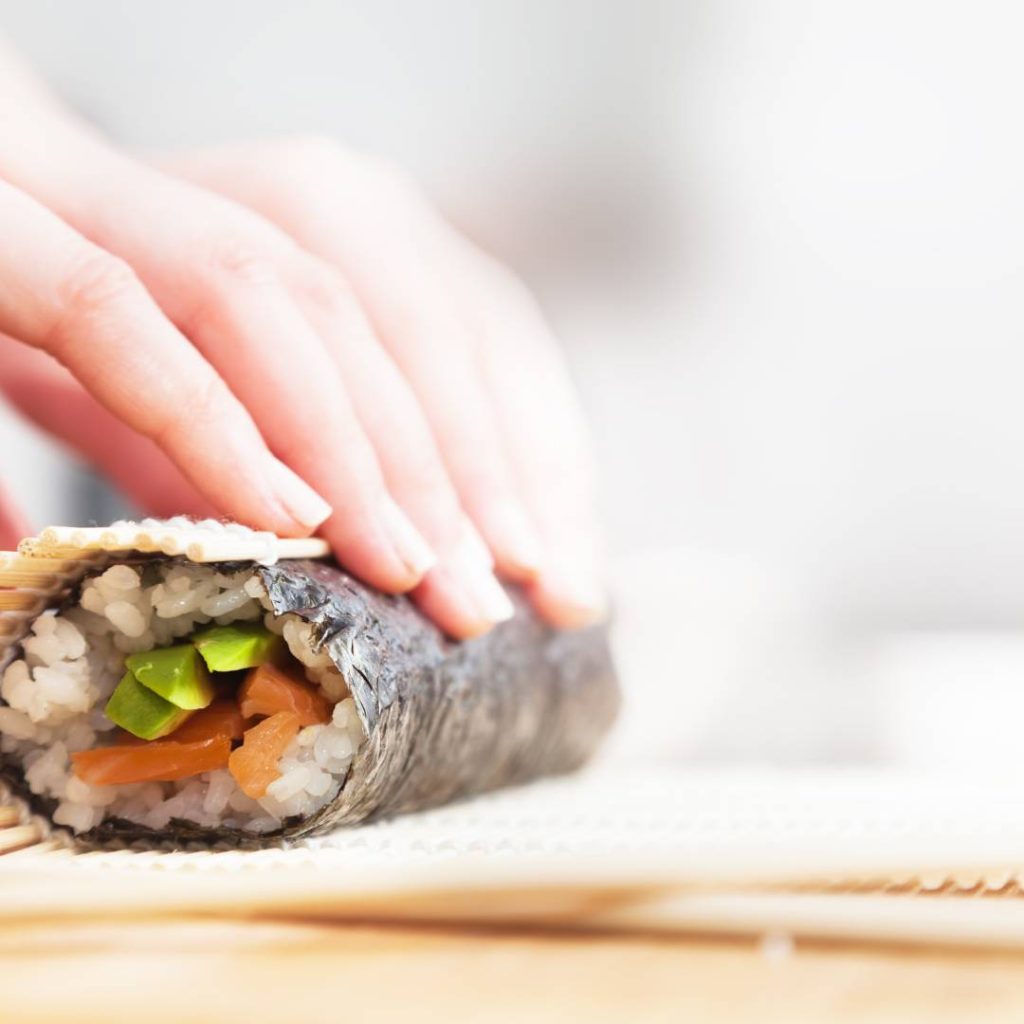 A woman's hand is preparing a sushi roll, focusing on the carbs in sushi.