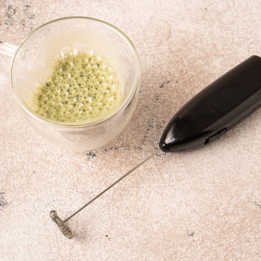 An electric whisk next to a cup of green smoothie.