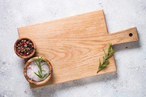 A wooden cutting board with salt, pepper and rosemary on a gray background.