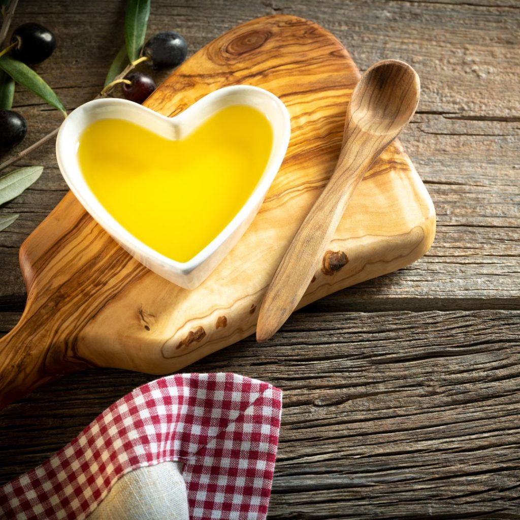 A heart container filled with olive oil lies on the table with fresh olives and wooden kitchen tools.