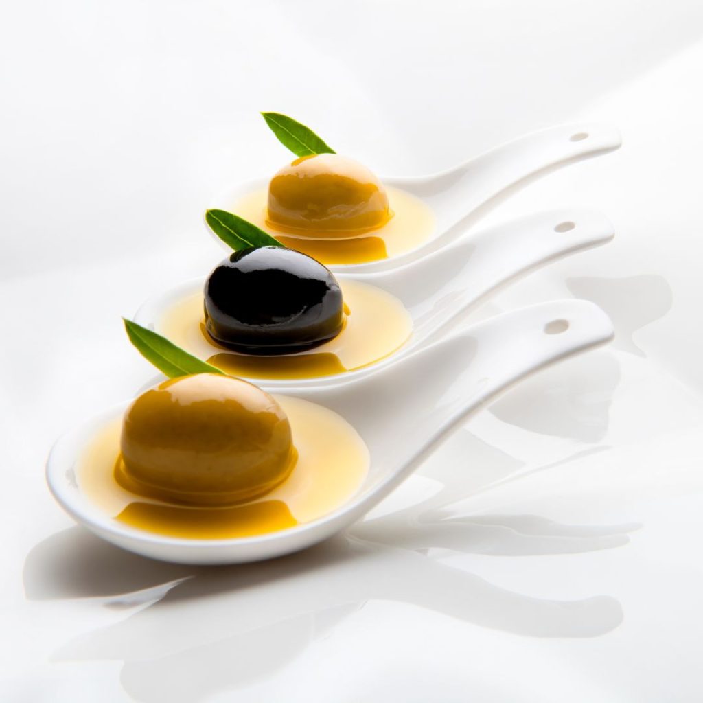 A line of olive varieties and olive oils rest on white spoons.