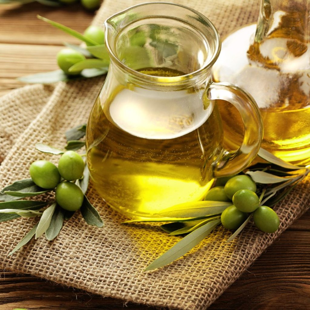 A pitcher of olive oil sits on the table with freshly picked olives.