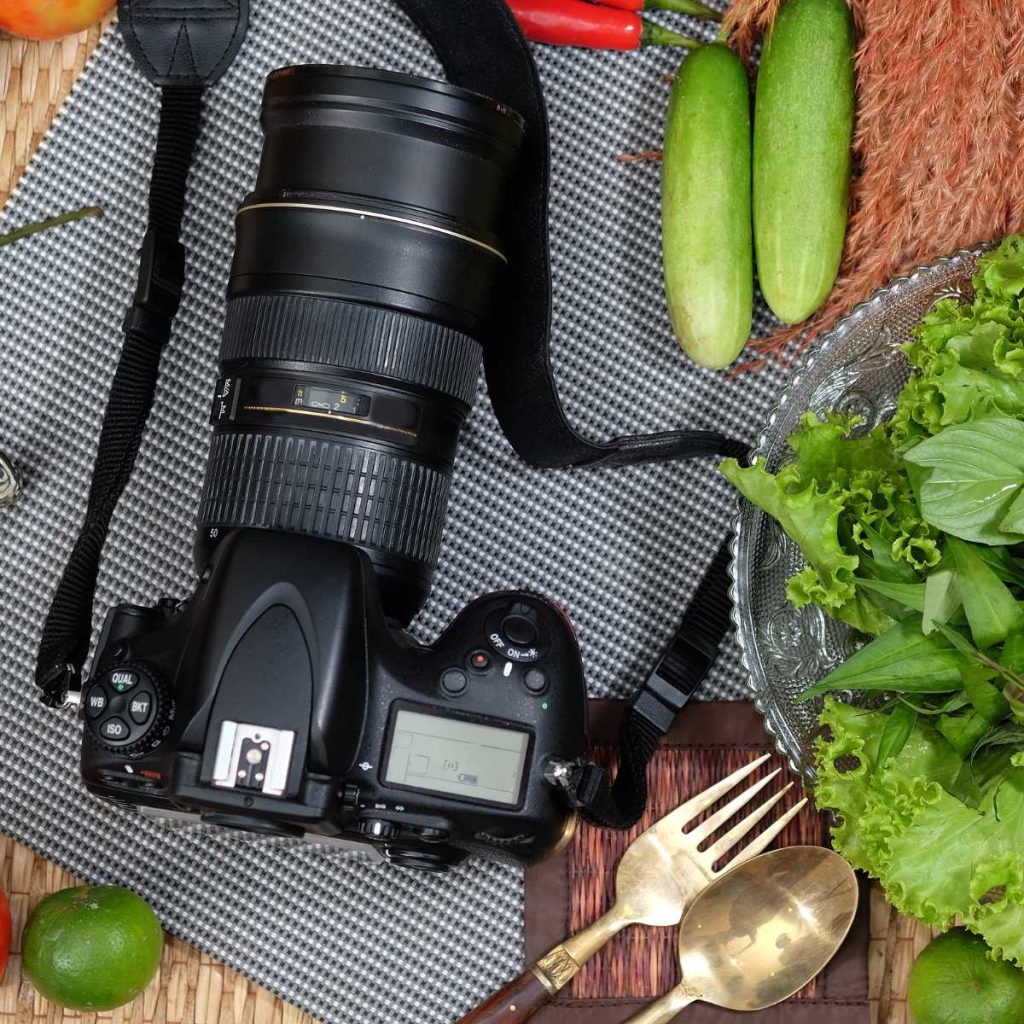 A camera with long focal length on a gray mat surrounded by vegetables and utensils for food photography