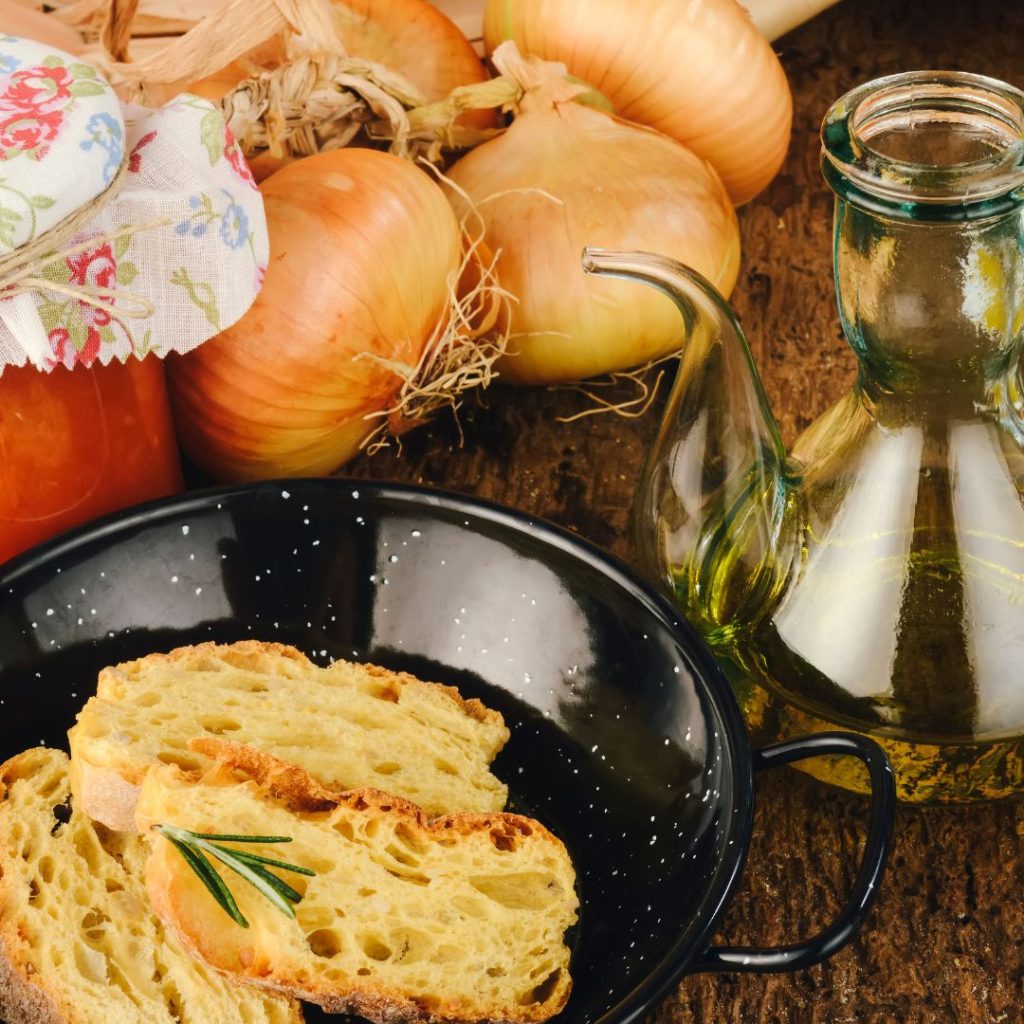 A bottle of olive oil sits on a wooden table with onions and toasts.