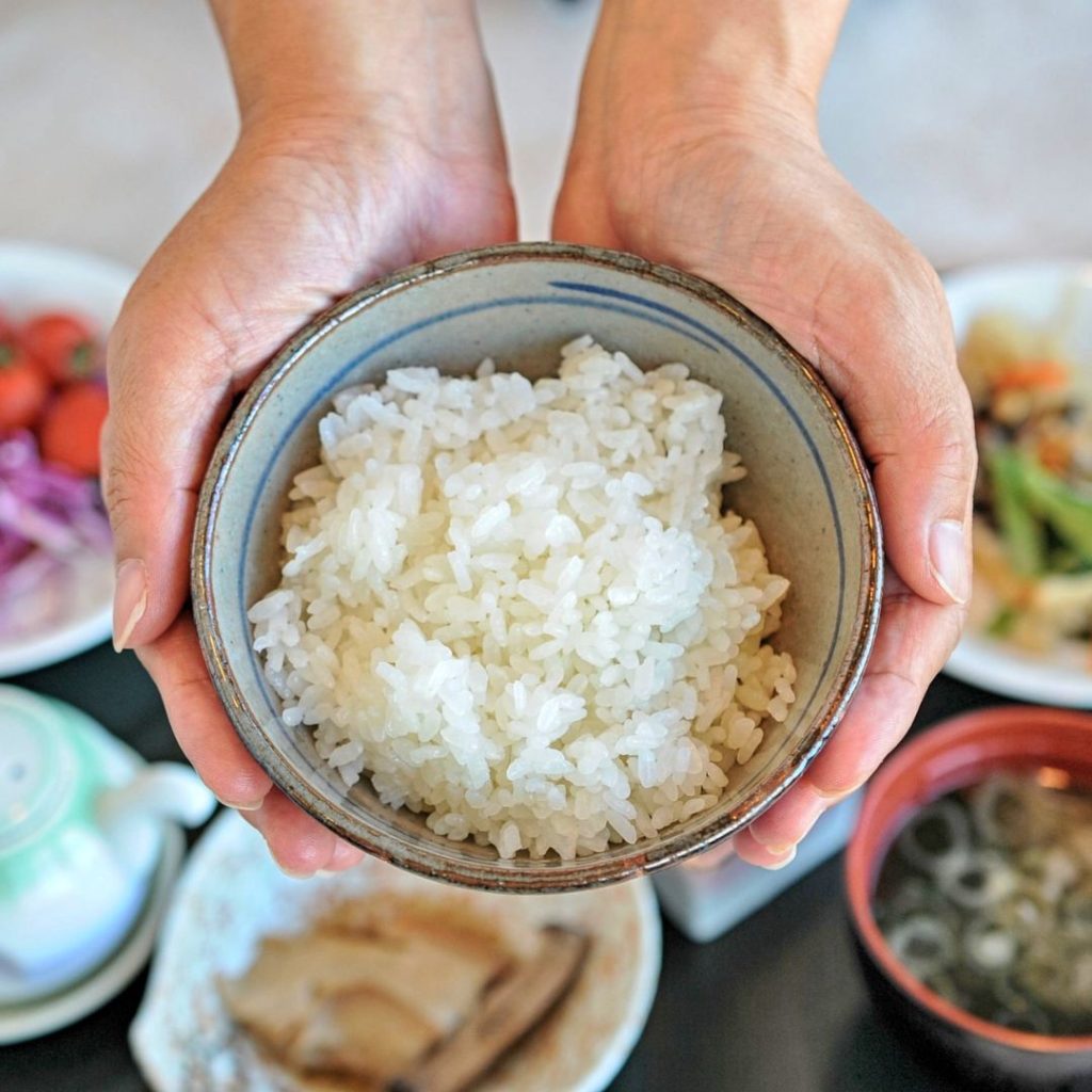 Two palms hold a plain rice bowl above other Japanese breakfast dishes.