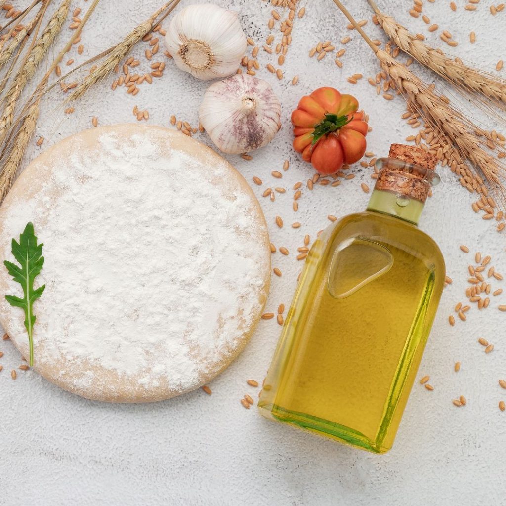 A bottle of extra virgin olive oil rests on a table with dough, garlic bulbs, wheat, and tomatoes.