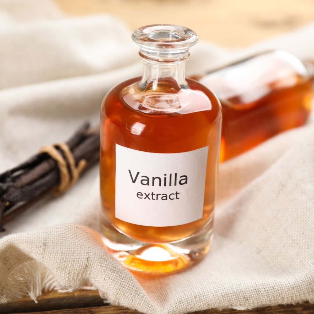 A bottle of vanilla extract rests on a tablecloth with some vanilla beans.