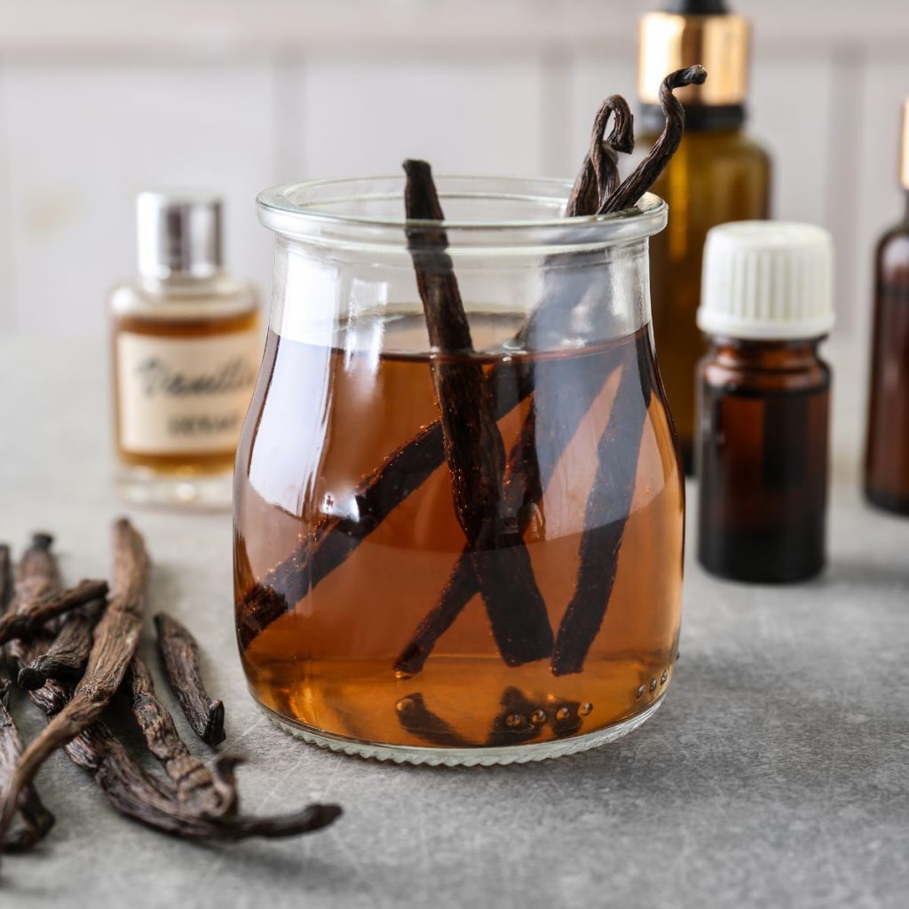 A container of vanilla extract rests on a tablecloth with some vanilla beans.