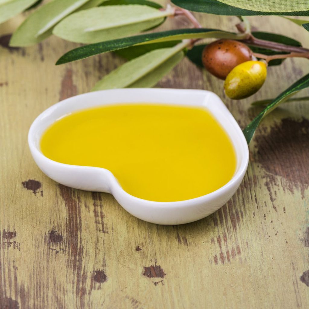 A heart-shaped container that contains extra virgin olive oil rests on a wooden table with some olives.