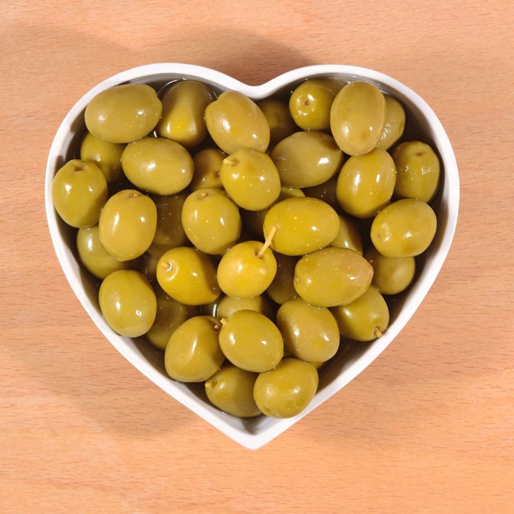 A heart-shaped ramekin is filled with olives soaked in extra virgin olive oil.