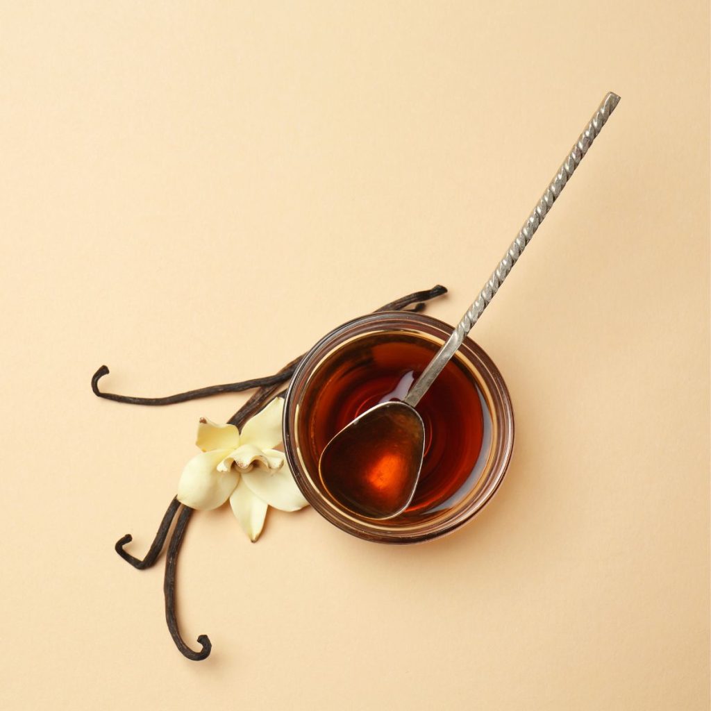 A ramekin full of vanilla extract rests on a pinkish table with some vanilla beans.