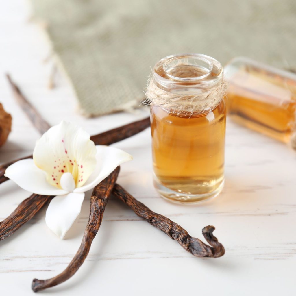 A small bottle of vanilla extract rests on a table with a set of vanilla beans and a vanilla orchid.