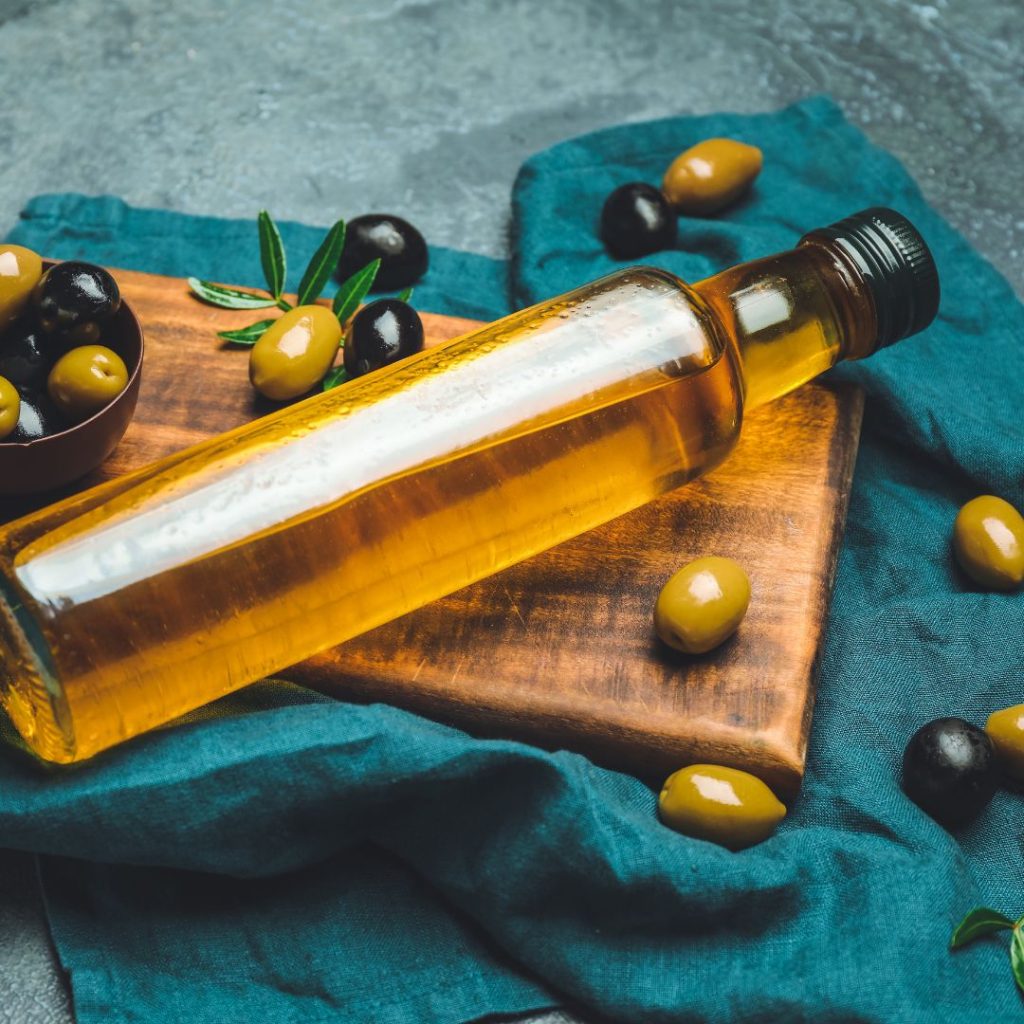 A bottle of olive oil rests on a wooden board and a cloth with olives.