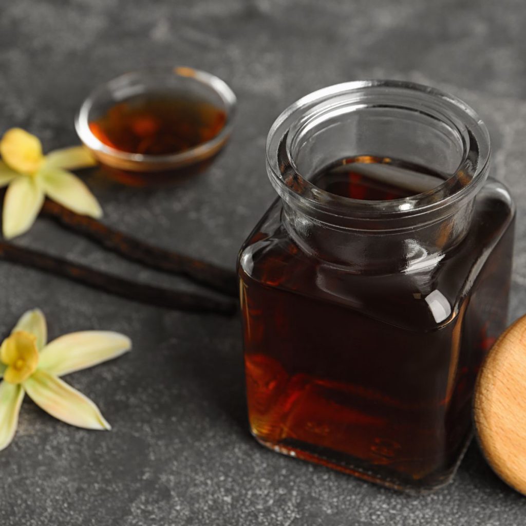 A container of vanilla extract rests on a table with some vanilla beans and a vanilla orchid.