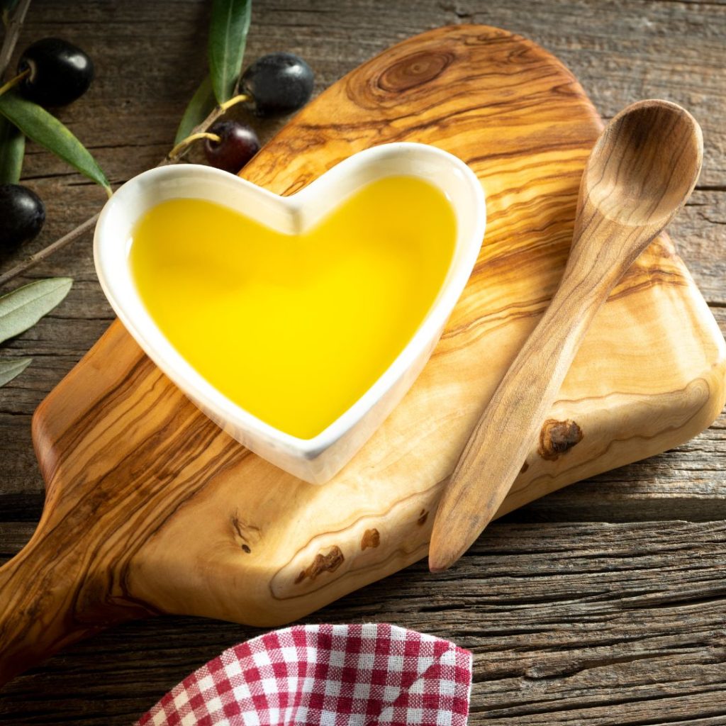 A heart-shaped ramekin full of extra virgin olive oil rests on a wooden board with a wooden spoon and some olives.