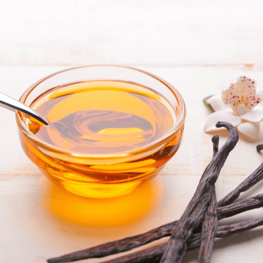 A sauce bowl of vanilla extract rests on a table with vanilla beans and vanilla orchids.