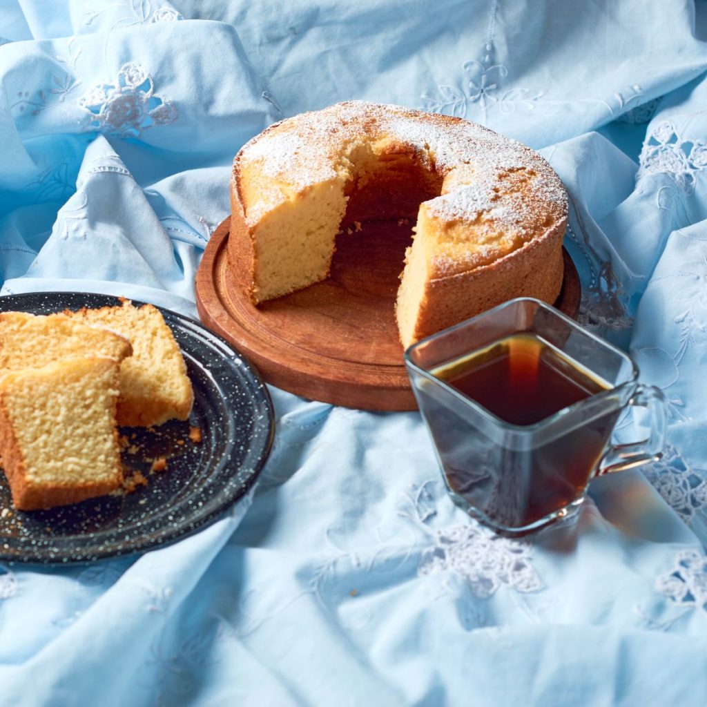 A small cup of vanilla extract rests on a tablecloth with two cakes.