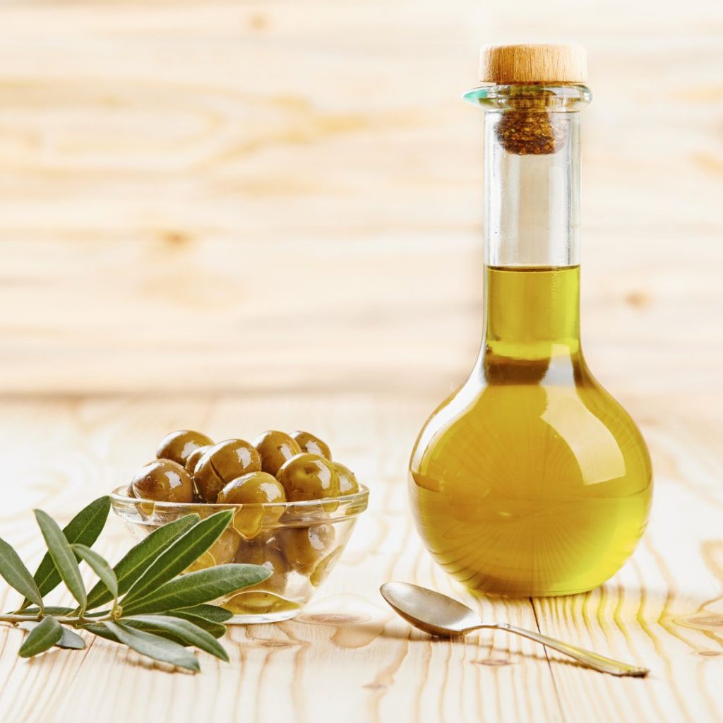 A bottle of extra virgin olive oil rests on a table with an ingredient bowl of olives.