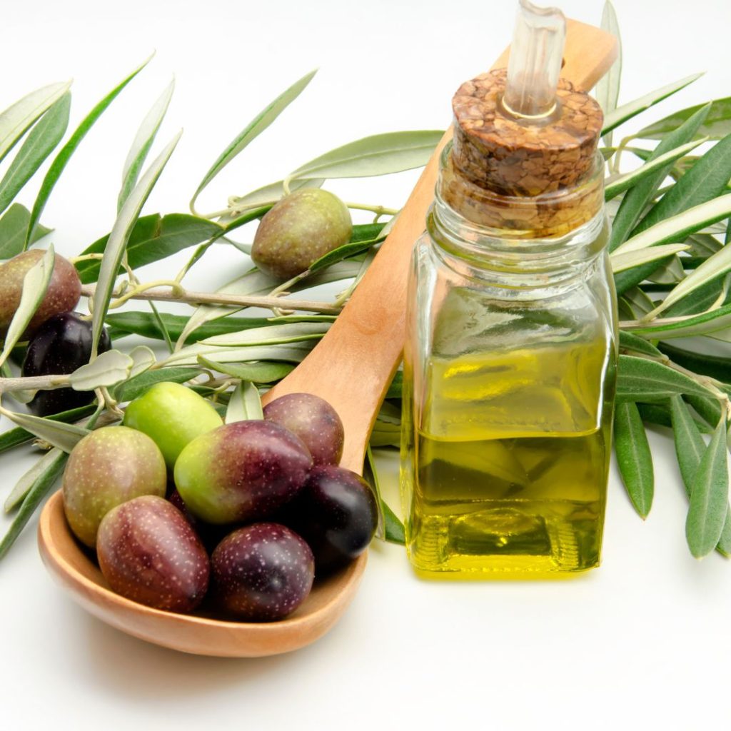 A bottle of extra virgin olive oil rests on a white table with a ladle full of olives.