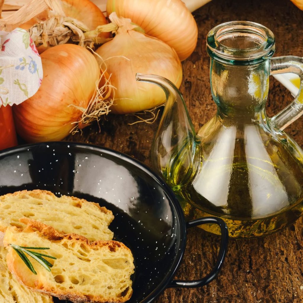 A pitcher of extra virgin olive oil, a bowl of toasted bread, and bulbs of onion rest on a wooden table.