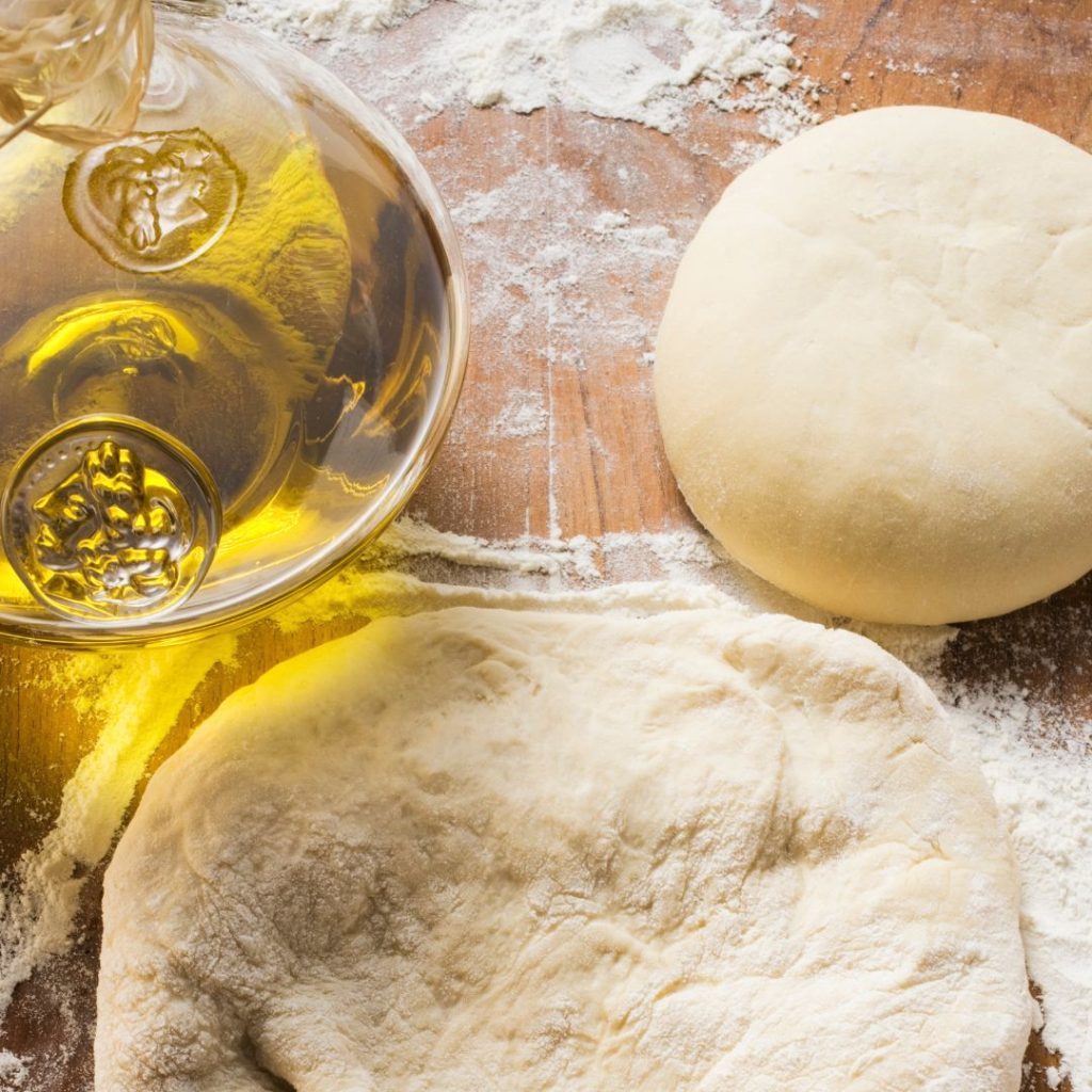 A pitcher of extra virgin olive oil rests on a table with a pair of dough.