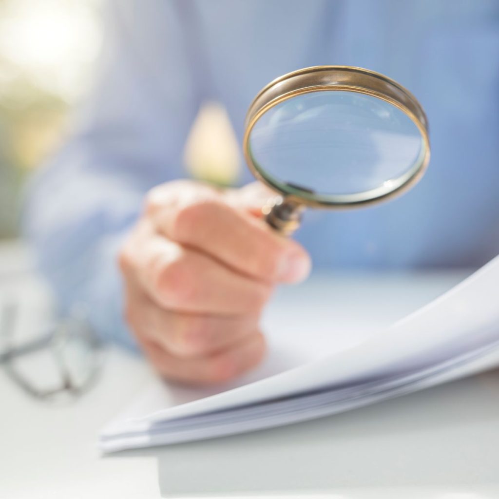 A hand holds a magnifying glass in front of papers.