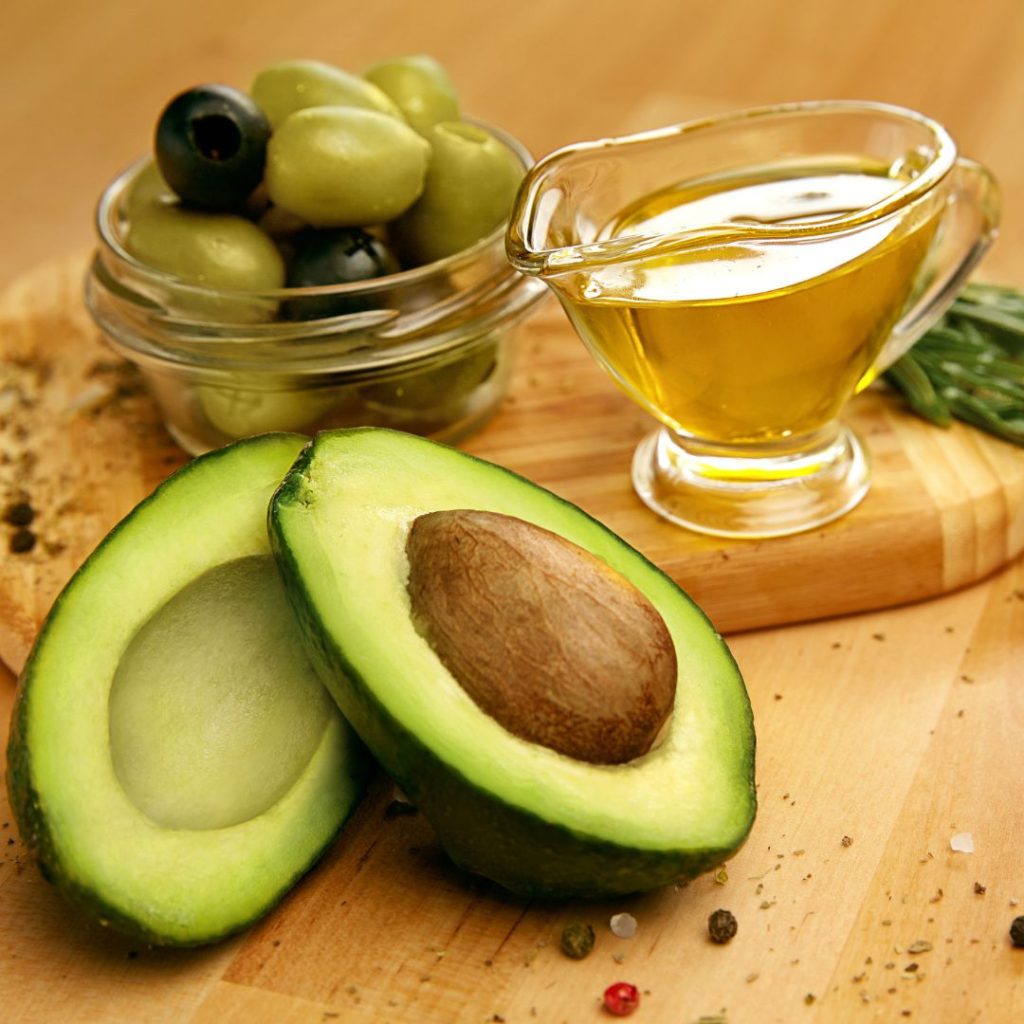 Avocados, olives, herbs, spices, and oil rest on a wooden board.