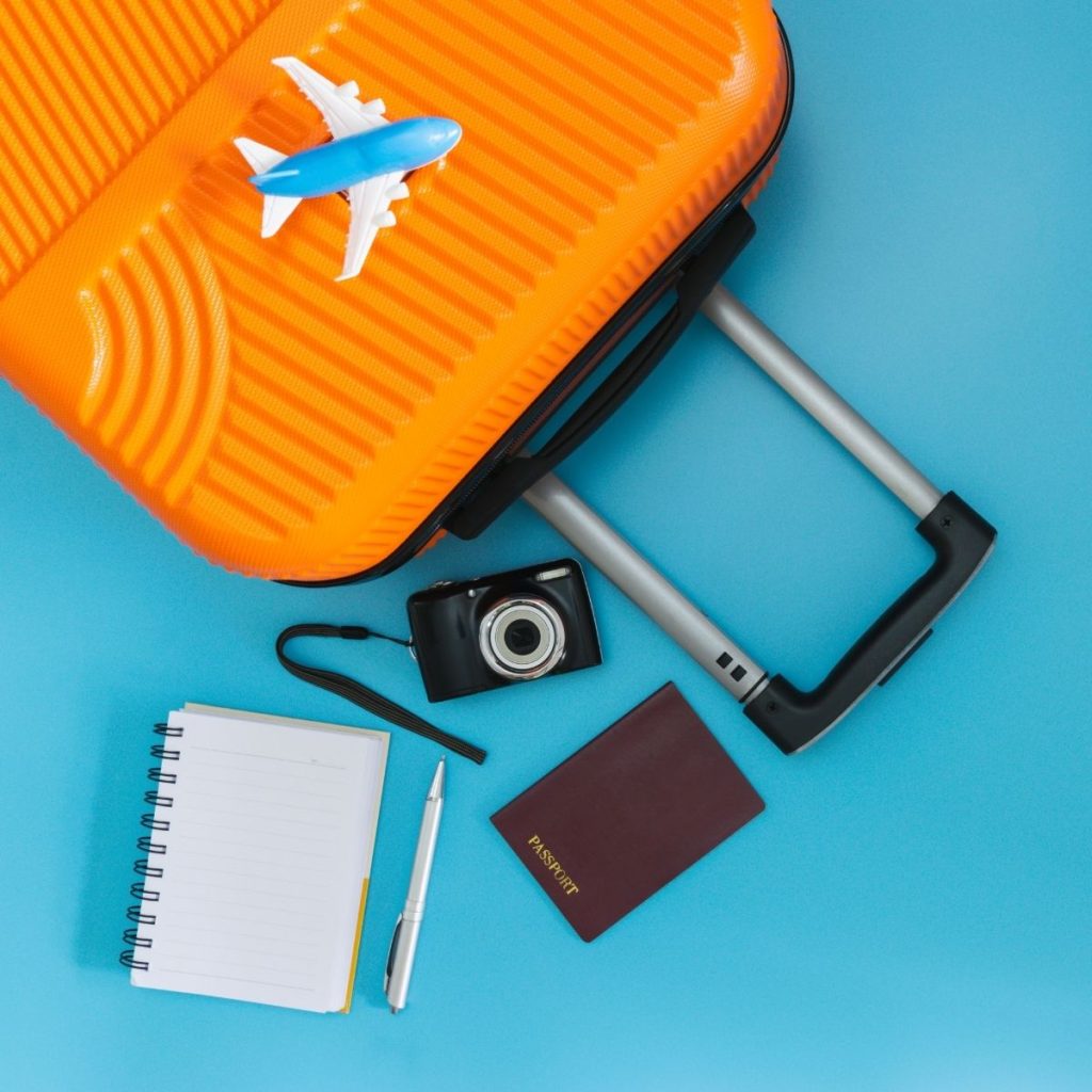 A luggage, notebook, pen, passport, and camera rest on a blue surface.