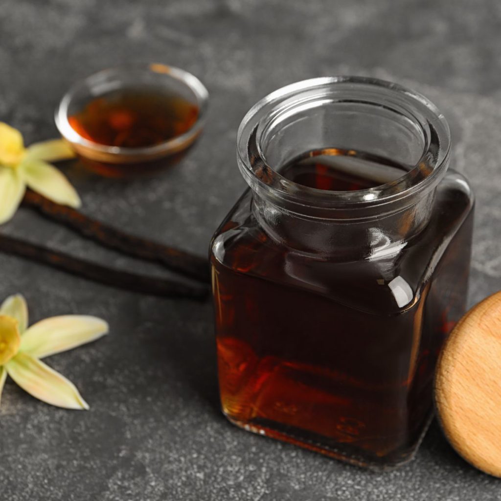 A small container of vanilla extract rests on a table with some vanilla beans and orchids.