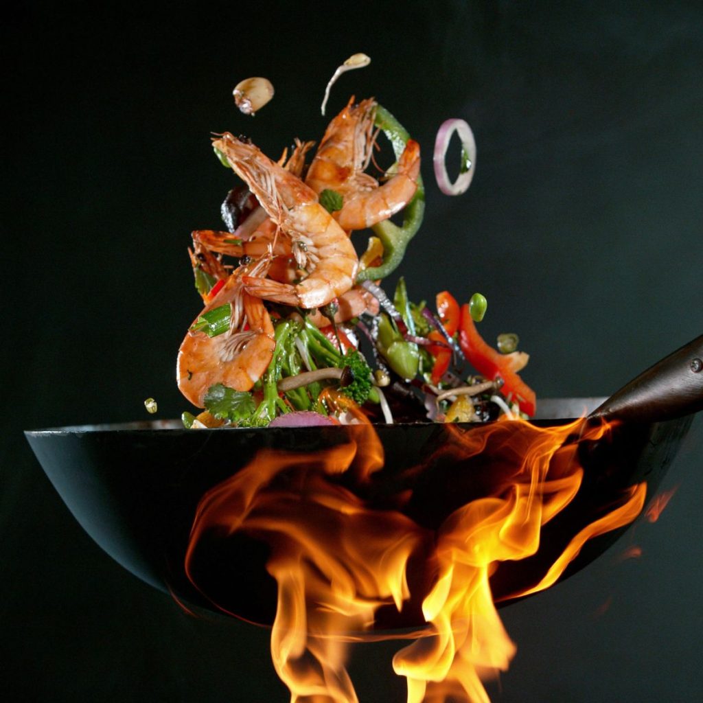 Seafood and vegetables are tossed with a metal wok under a fire.