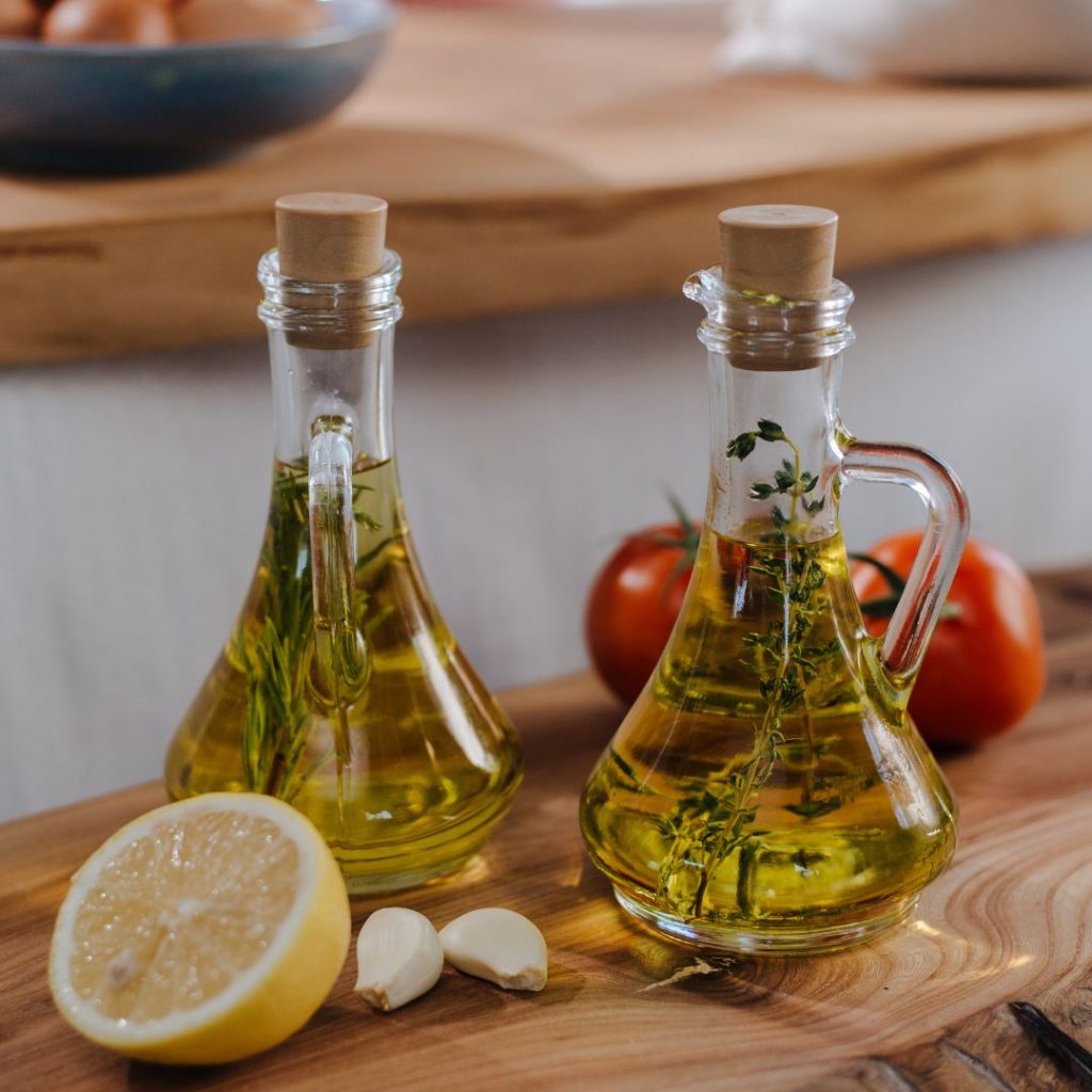 Two bottles of extra virgin olive oil rest on a wooden table with a lemon, cloves of garlic, and tomatoes.