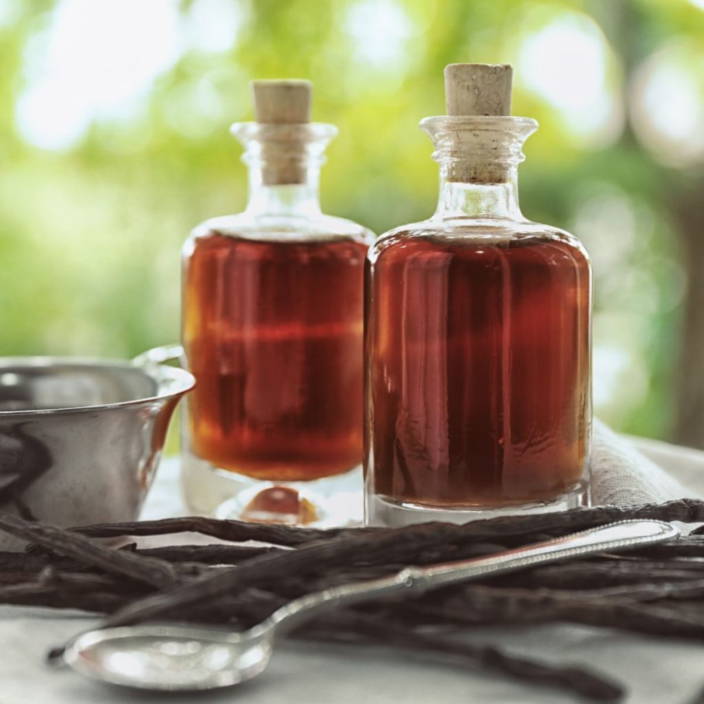 Two bottles of vanilla extract rest on a table with a spoon and some vanilla beans.