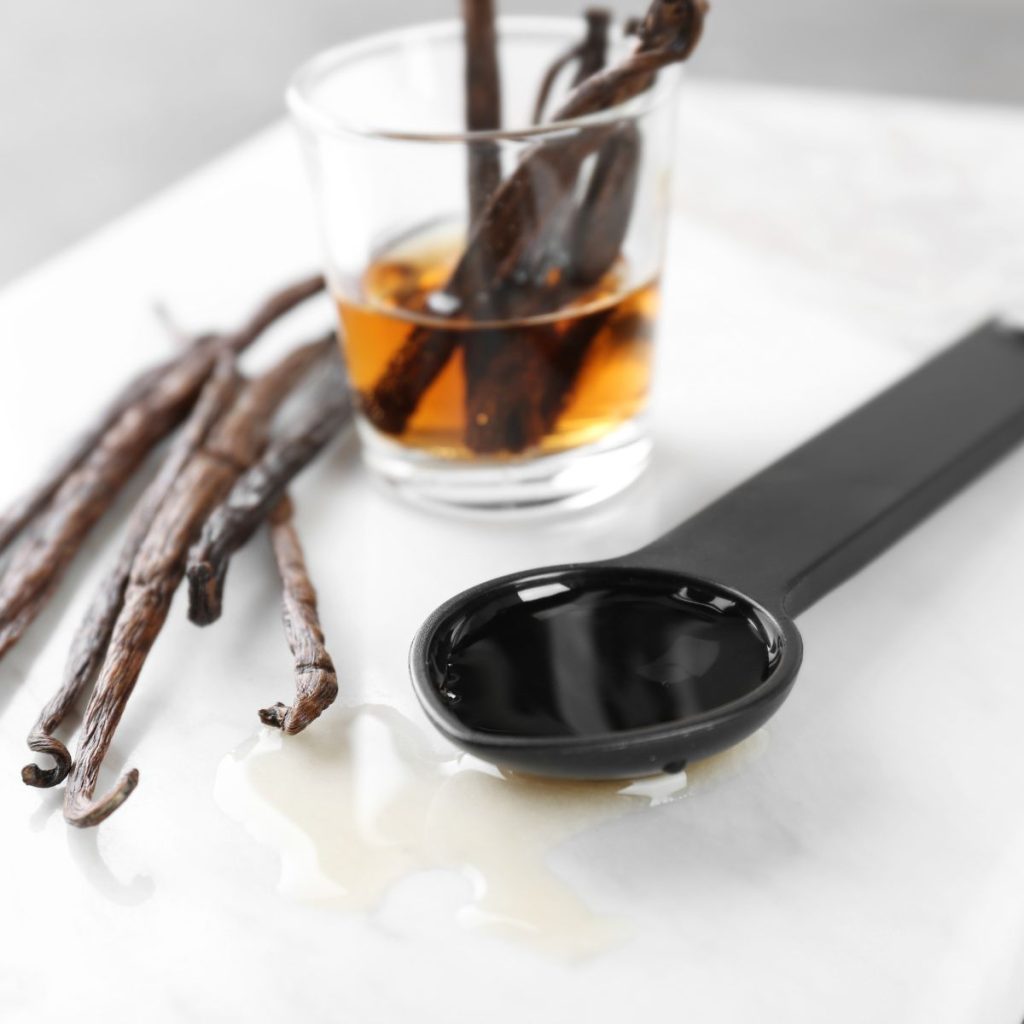 A small cup and a spoonful of vanilla extract rest on a white table with some vanilla beans.