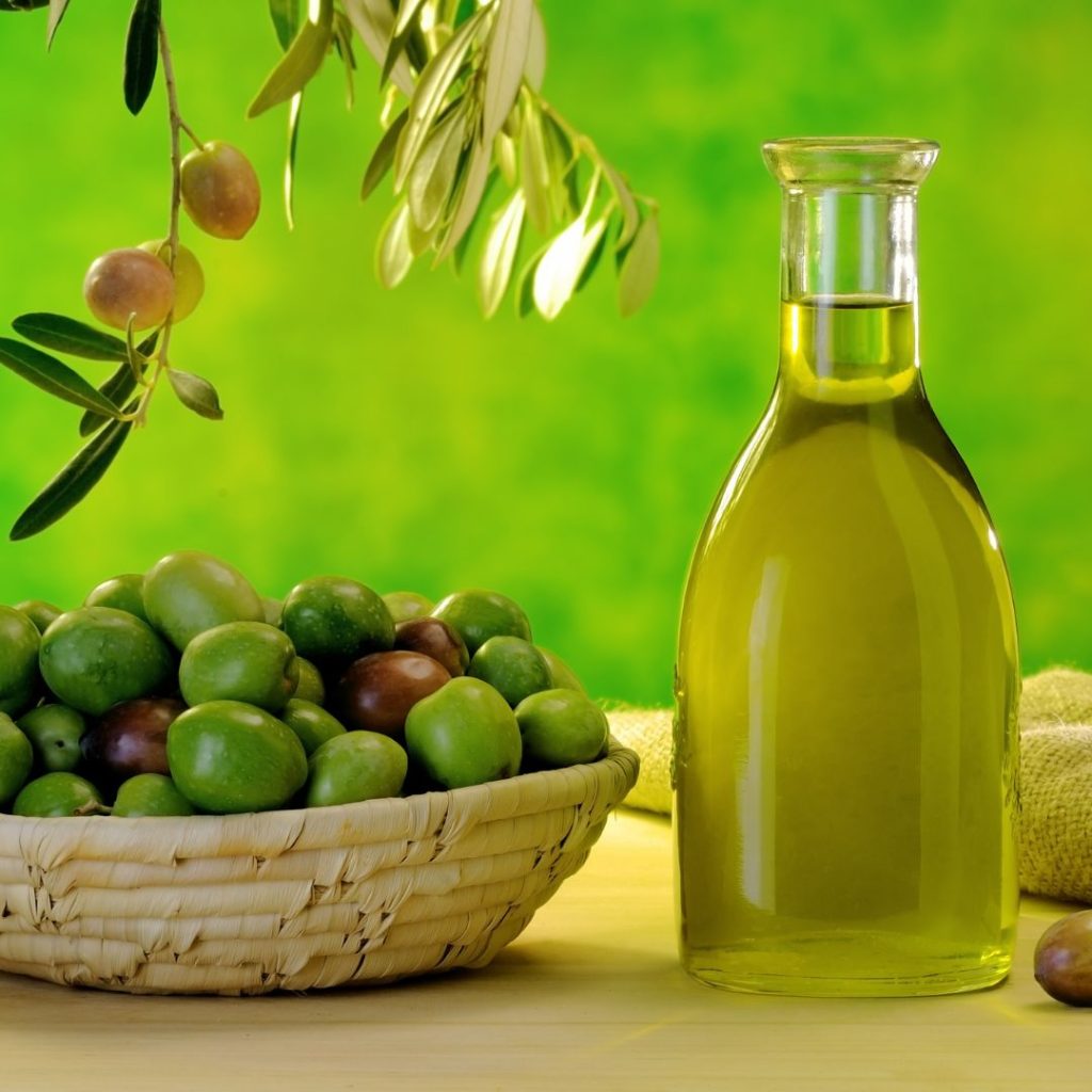 A bottle of extra virgin olive oil rests on a table with a basket of olives.