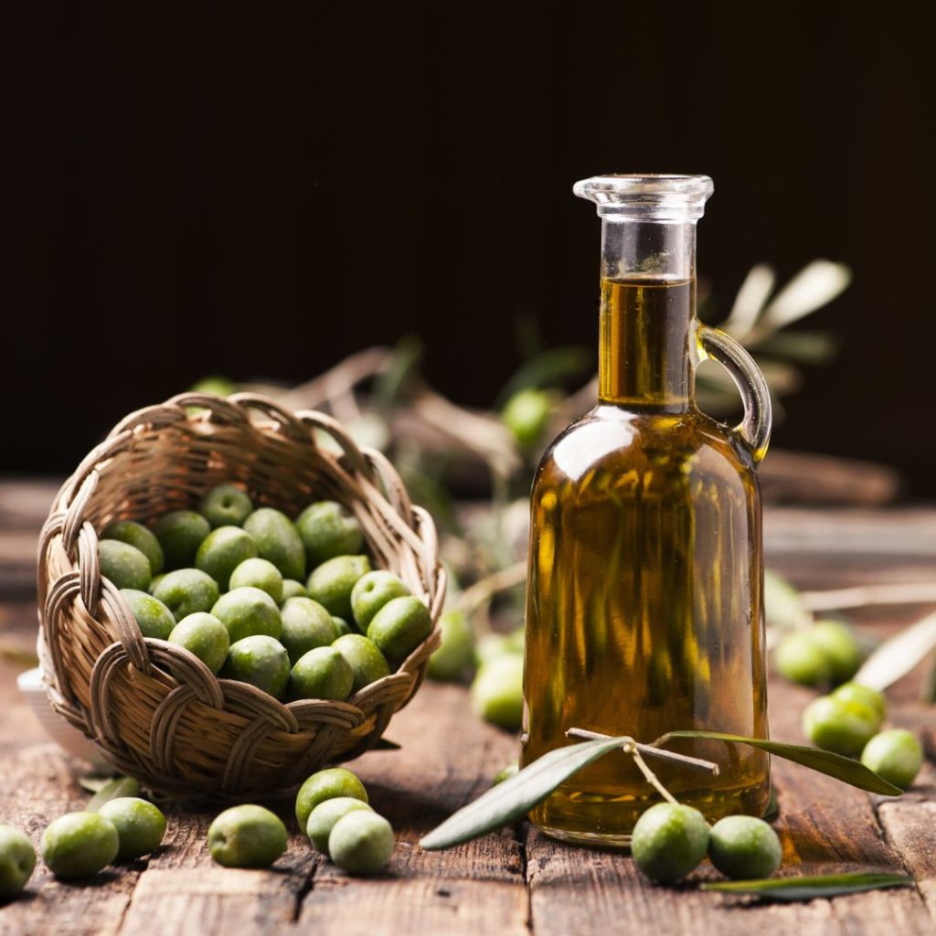 A bottle of extra virgin olive oil sits on a table with a basket of olives.